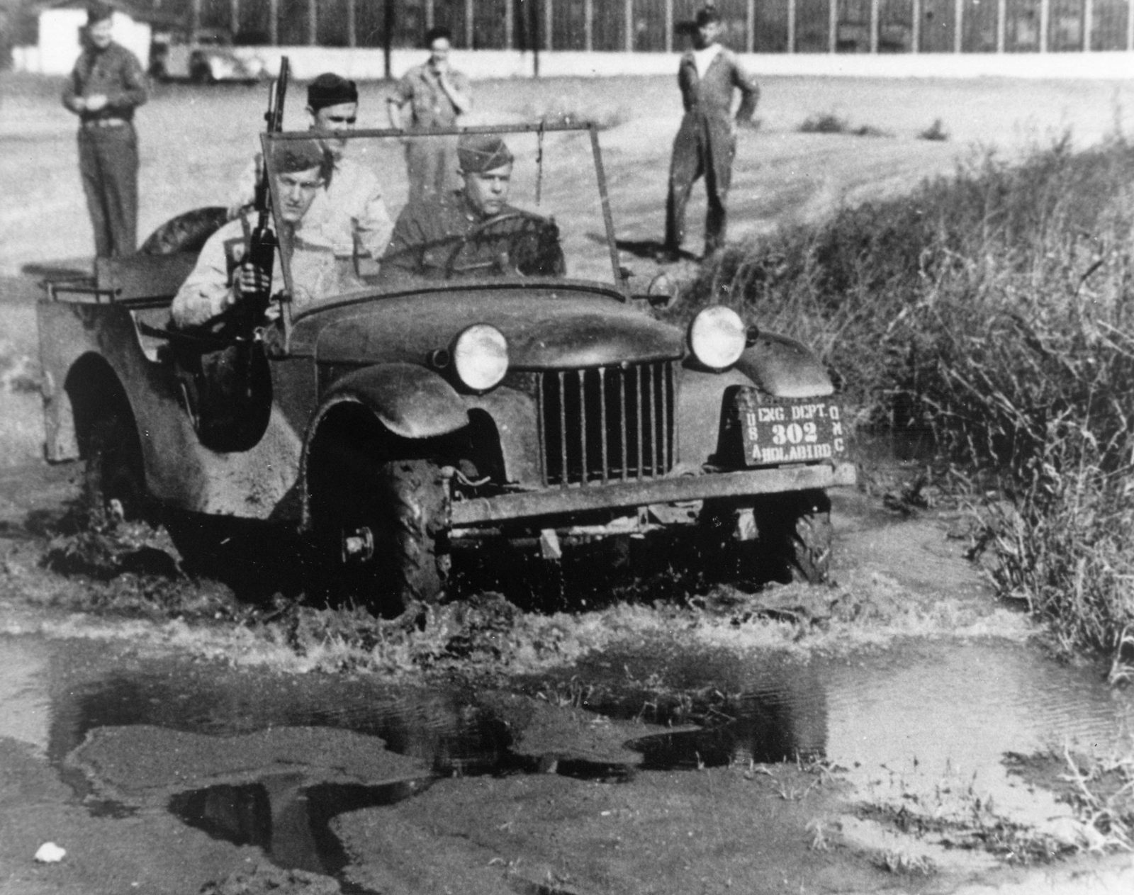 The Bantam pilot model, a small open car with rounded fenders, driving through mud and water with three soldiers aboard.