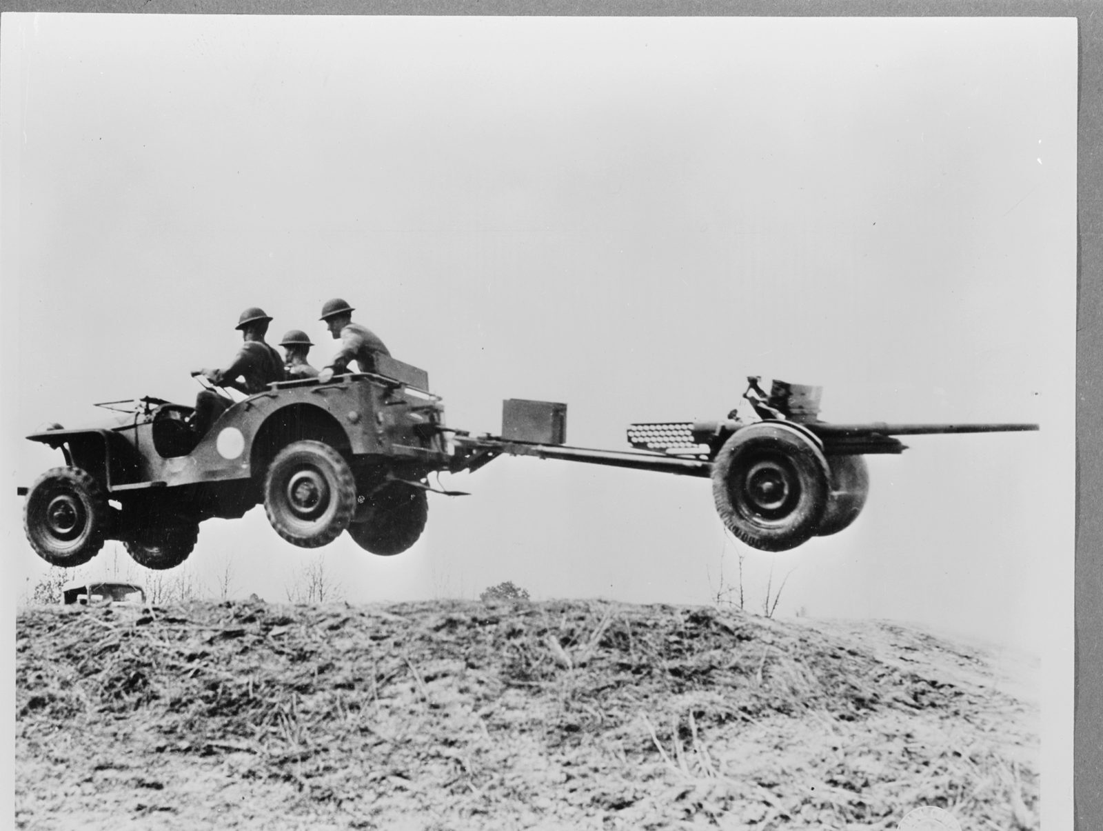 A Bantam with three soldiers aboard leaps over a rise with all four wheels off the ground, towing a 37 mm anti-tank gun.